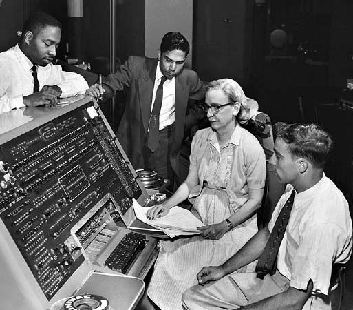 Grace Hopper at the control panel/console of the UNIVAC Computer, surrounded by coworkers looking on